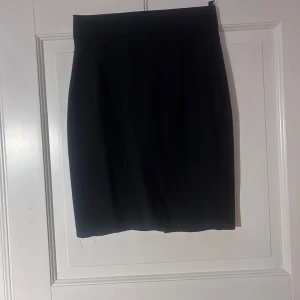 Svart kortkjol med slits, stl 38 - Enkel och stilren svart kortkjol i klassisk modell. Kjolen har en liten slits framtill som ger en snygg detalj. Passar perfekt till en trendig look och är lätt att matcha med olika toppar och jackor.