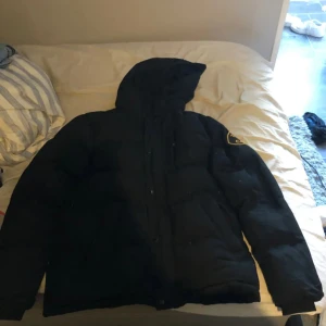 Svart pufferjacka från Zavetti Canada - Säljer en svart pufferjacka från Zavetti Canada med huva och patch på ärmen. Jackan är rejält vadderad och har en clean look med dragkedja och knappar framtill. Perfekt för kalla dagar när du vill hålla stilen och värmen. Den är lite nopprig har ett hål på armen som inte syns.