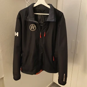 Svart vindjacka Helly Hansen - Svart vindjacka från Helly Hansen med marinblå detaljer och vita loggor på bröstet, ryggen och ärmen. Jackan har dragkedja framtill, röda dragkedjor på fickorna och är tillverkad i slitstark polyester. Perfekt för dig som gillar en sportig och nautisk stil.