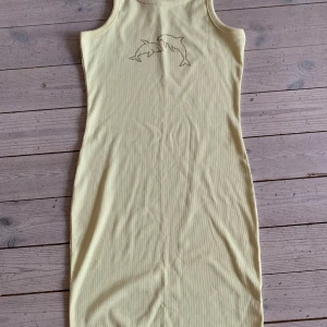 Gul ribbad klänning med delfiner Lindex - Denna klänning har använts fåtal gånger. Söt ljusgul ribbad klänning från Lindex med tight passform. Ärmlös modell med rund halsringning och glittriga delfiner framtill. Perfekt för sommarens varma dagar och enkel att styla med sneakers eller sandaler. 