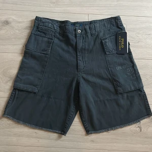 Svarta cargoshorts från Polo Ralph Lauren - Svarta cargoshorts från Polo Ralph Lauren med stora fickor på sidorna och råa kanter nertill. Klassisk midja och tillverkade i mjuk bomull. Snygg, avslappnad stil med diskreta tryckdetaljer på ena benet. Perfekta för dig som gillar streetwear och praktiska detaljer.