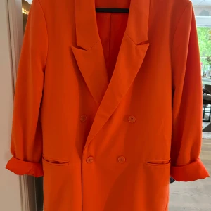 Orange kavaj med dubbelknäppning - Snygg orange kavaj med dubbelknäppning och klassisk krage. Jackan har långa ärmar med uppvikta muddar och två fickor framtill. Tillverkad i en mix av polyester, viskos och elastan för en skön och följsam känsla.