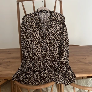 Leopardmönstrad klänning från Zara - Leopardmönstrad kortklänning från Zara Woman med volang nertill, långa ärmar med knapp vid ärmslut och knytning i halsen. Klänningen har beige och svarta toner och är tillverkad i ett lätt, flowy material som ger en snygg siluett.