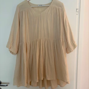 Beige trekvartsärmad klänning från NA-KD - Luftig och flowy otroligt mjuk minklänning från NA-KD i beige. Klänningen har v-ringning, trekvartsärmar och flera volanger. Aldrig använd, därmed mycket fint skick🤍