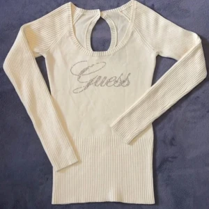 Vintage tröja med strass från Guess - Supersnygg vit stickad tröja från Guess med glittrande strasslogga framtill. Tröjan har långärmade ribbade ärmar, bred ribbad nederkant och en öppen detalj i ryggen som ger en unik look. Perfekt för dig som gillar stilrena men ändå iögonfallande plagg.
