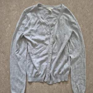 Kofta - Ljusgrå kofta från H&M Basic i storlek XS. Klassisk modell med rund halsringning och knäppning framtill med små knappar. Mjuk och tunn, perfekt att slänga över en topp. Ribbstickade muddar vid ärmslut och nederkant. Enkel och stilren för alla tillfällen.