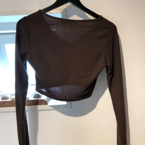 Brun ribbad croppad topp Boohoo - Säljer en brun croppad långärmad topp från Boohoo. Toppen är ribbad med v-ringning och har en tight passform. Perfekt att styla med höga jeans eller kjol för en trendig look.