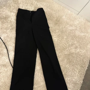 Svarta kostymbyxor med raka ben - Klassiska svarta kostymbyxor med raka ben och hög midja. Byxorna har hällor för bälte och stängs med dragkedja framtill. Perfekta för en clean och stilren look.