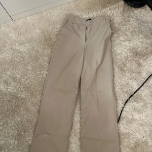 Beige kostymbyxor med raka ben - Stilrena beige kostymbyxor med raka ben och hällor för bälte. Klassisk design med diskret dragkedja framtill. Perfekta för en clean och dressad look.