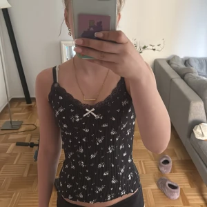 Svart blommig linnetopp från H&M Divided - Supersöt svart linnetopp med vita små blommor och tunna ränder från H&M Divided. Toppen har spetskant upptill, justerbara axelband och en liten rosett framtill. Figurnära passform och croppad längd, perfekt till jeans eller kjol.