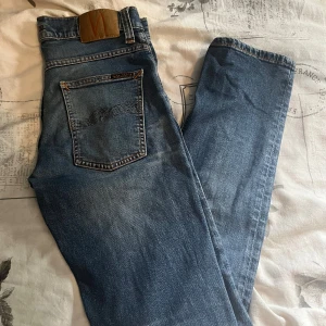 Nudie Jeans - Säljer ett par klassiska blå jeans från Nudie Jeans med snygg tvätt och kontrastsömmar. Jeansen har rak passform och är tillverkade i bomull. Perfekta för dig som gillar en tidlös och avslappnad stil.