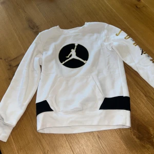 Vit Jordan tröja med guldtryck - Snygg vit Jordan tröja med svart och guld logga på bröstet och guldtext på ärmen. Tröjan har svarta detaljer vid midjan och en stor ficka framtill. Långärmad och i mjuk bomull, perfekt för en sportig look.