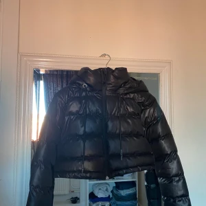 Svart croppad pufferjacka Pickk Upp - Säljer en svart croppad pufferjacka från Pickk Upp. Jackan har huva med snörning, dragkedja framtill och en glansig finish. Perfekt för dig som vill ha en trendig och varm jacka med cool attityd.