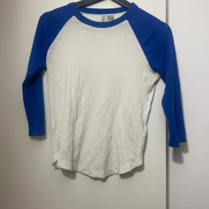 Vit och blå långärmad tröja H&M - Vit långärmad tröja från H&M Divided med blå ärmar och blå krage. Klassisk baseballstil med rund halsringning och normal passform. Tillverkad i mjuk bomull, perfekt för en chill och sportig look.