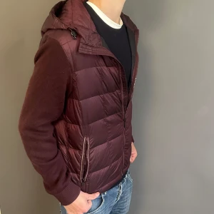 Vinröd pufferjacka från Massimo Dutti - Snygg vinröd pufferjacka från Massimo Dutti med stickade ärmar och vadderad kropp. Jackan har huva, dragkedja framtill och två fickor med dragkedja. Perfekt för dig som vill ha en stilren och bekväm jacka med modern känsla.