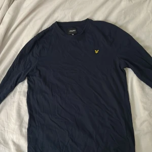 Mörkblå långärmad tröja Lyle & Scott - Snygg mörkblå långärmad tröja från Lyle & Scott med klassisk gul logga på bröstet. Enkel och stilren design, perfekt till jeans eller chinos. Tillverkad i mjuk bomull som känns skön mot huden.