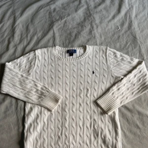 Vit kabelstickad tröja Polo Ralph Lauren - Vit kabelstickad tröja från Polo Ralph Lauren med klassisk rund halsringning och broderad logga på bröstet. Tröjan har långa ärmar och ribbade muddar, perfekt för en clean och tidlös stil. Passar från 165 till 175 och priset kan diskuteras vid snabb affär