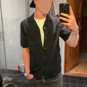 Svart kortärmad skjorta - Snygg svart skjorta med kort ärm och klassisk krage. Skjortan har knappar framtill och en bröstficka, perfekt att bära öppen över en t-shirt. Enkel och stilren design som passar till många outfits.