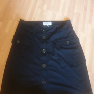 Svart kjol med knappar från Freequent - Svart kjol från Freequent med hög midja, stora knappar framtill och två fickor med lock. Kjolen har bälteshällor och är i en klassisk, rak modell som slutar ovanför knät. Perfekt för dig som gillar stilrena och enkla plagg.