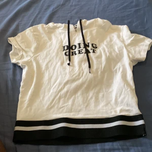 Vit hoodie med tryck från FB Sister - Vit hoodie från FB Sister med svart text 'DOING GREAT' på bröstet. Kortärmad modell med svart-vita ränder längst ner och snörning i huvan. Skön och avslappnad passform, perfekt för en chill look.