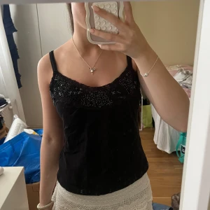 Svart linnetopp med pärlbroderi - Supersnygg svart linnetopp med smala axelband och dekorativt pärlbroderi upptill. Toppen har en lätt glansig yta och spetsdetaljer längs urringningen som ger en lyxig känsla. Perfekt att styla med kjol eller jeans för en trendig look.