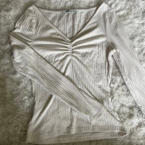 Ribbad v-ringad topp från NA-KD - Säljer en ljusbeige ribbad topp från NA-KD med v-ringning och snygg rynkdetalj framtill. Långärmad och figurnära passform, perfekt till jeans eller kjol. Superenkel att styla och passar året runt. Liten fläck på insidan av ryggen, går inte bort i tvätt. Syns ej när tröjan är på.