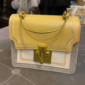 Gul och vit handväska med gulddetaljer - Snygg handväska i gul och vit med coolt guldhandtag och lås framtill. Väskan har en boxig form, kontrasterande sömmar och detaljer i skinn och textil. Perfekt för dig som vill sticka ut med accessoarer som känns unika och trendiga.