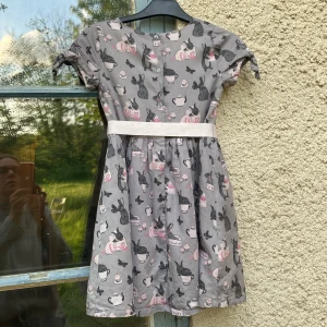 Grå kaninklänning med rosett från Lindex - Grå  kortklänning från Lindex med gulligt kaninmönster, tekannor och cupcakes i svart och rosa. Klänningen har korta ärmar med knyt, rund hals och en ljusrosa midjerem med rosett.   Rosa foder och underkjol i luftigt material.