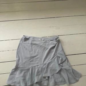 Ljusgrå/blå kort volangkjol  - Säljer en superfin ljusgrå kjol från Nakd. Den har en snygg volangdetalj som ger en extra touch av stil. Perfekt för både vardag och fest! Kjolen är i storlek 40 och är i bra skick. Passar perfekt till en somrig dag eller en mysig höstkväll. 🌟