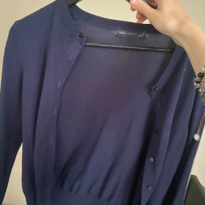 Mörkblå kofta från M&S Collection - Säljer en klassisk mörkblå kofta från M&S Collection. Koftan har rund halsringning, långa ärmar och knäppning framtill med matchande knappar. Perfekt att slänga över en topp eller klänning.