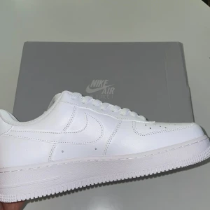 Nike Air Force 1 helvita sneakers - Klassiska helvita Nike Air Force 1 sneakers med rund tå och platt sula. Skorna har snörning och är tillverkade i skinn. Perfekta för dig som gillar en clean och tidlös stil.