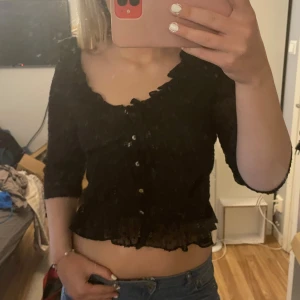 Svart croppad blus med volang och knappar - Säljer en svart croppad blus med volangkant och knappar framtill. Blusen har trekvartsärm och lätt transparent material med diskret prickigt mönster. Perfekt för dig som vill ha en söt och trendig topp med fina detaljer.
