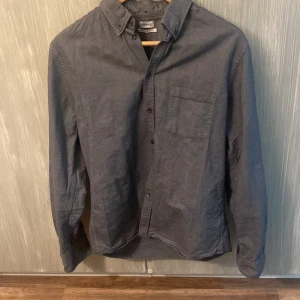 Grå skjorta från Filippa K - Stilren mörkgrå skjorta från Filippa K i storlek Small. Skjortan har klassisk krage med knappar, bröstficka och långa ärmar. Perfekt för en clean och enkel look. Tillverkad i mjukt bomullstyg.