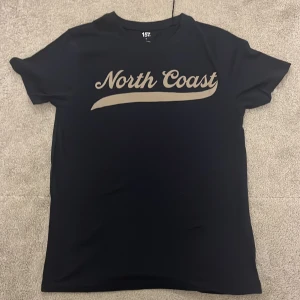 Svart t-shirt med tryck från 157 - Svart t-shirt från 157 med beige texten 'North Coast' tryckt på bröstet. Klassisk passform och rund halsringning. Perfekt för en avslappnad stil. Jag köpte den för 60kr och den var för lite och jag tappade bort kvittot