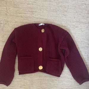 Vinröd stickad cardigan jacka med stora knappar - Mysig vinröd stickad cardigan jacka med stora guldiga knappar framtill och två fickor. Jackan har rund halsringning och lång ärm, perfekt för lager på lager under kyliga dagar. Den är i storlek M men passar även S 