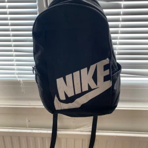 Svart ryggsäck från Nike - Säljer en svart ryggsäck från Nike med stort vitt logotryck på framsidan. Väskan har justerbara axelremmar och ett stort huvudfack samt ett mindre ytterfack med dragkedja. Perfekt för skola eller träning.
