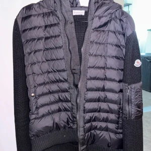 Moncler cardigan - Säljer en svart Moncler-jacka med quiltad framsida och huva. Ärmar och rygg är stickade vilket ger en snygg kontrast. Jackan har dragkedja och en ficka med dragkedja på ärmen. Perfekt för kyliga dagar. NFC-scan funkar. Strl L men passar M