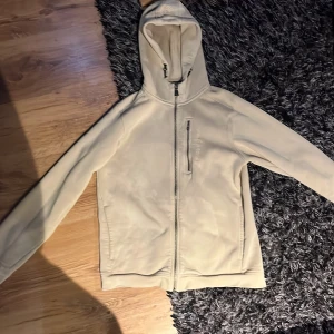 Beige fleecejacka med huva från Sail Racing - Beige fleecejacka från Sail Racing med huva och hel dragkedja framtill. Jackan har två sidofickor med dragkedja och en bröstficka med dragkedja. Mjuk insida i teddyfleece och broderad logga på bröstet. För liten för mig så de är därför jag vill få bort den, bröst fickan har dragkedjan gått sönder. Men den funkar fortfarande bara att man inte har det lilla taget att dra i