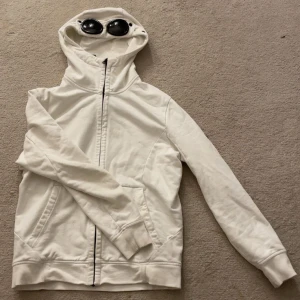 Vit hoodie med glasögon i huvan från C.P. Company - Säljer en vit hoodie från C.P. Company med unika inbyggda glasögon i huvan. Jackan har dragkedja framtill, två fickor och långa ärmar. Perfekt för dig som vill sticka ut med en ikonisk streetwear-stil.