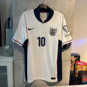 England fotbollströja Bellingham 10 - Säljer en vit England herrlandslagets fotbollströja med mörkblå detaljer, nummer 10 och Bellingham på ryggen. Tröjan har korta ärmar, broderad landslagslogga, Nike-logga och officiella turneringsmärken på ärmarna.