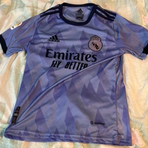 Real Madrid tröja - Säljer en blå/lilla Real Madrid bortatröja från Adidas med svart logga och klubbmärke på bröstet. Tröjan har korta ärmar, geometriskt mönster och svart krage. Emirates Fly Better som sponsortryck på framsidan.