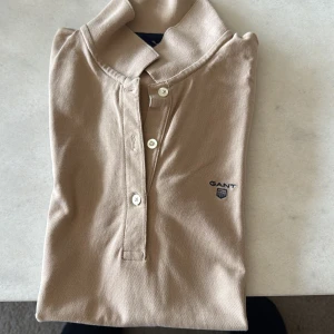 Beige pikétröja från GANT - Säljer en klassisk beige pikétröja från GANT med krage och knäppning framtill. Tröjan har diskret GANT-logga broderad på bröstet och är perfekt för en stilren look.