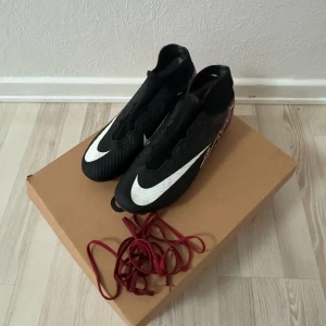 Nike fotbollsskor med svart och röd design - Säljer ett par Nike fotbollsskor i svart med stora vita swoosh-loggor och röda detaljer med AIR-tryck på sidorna. Skorna har högre skaft och röda dobbar undertill. Perfekta för dig som vill sticka ut på planen.