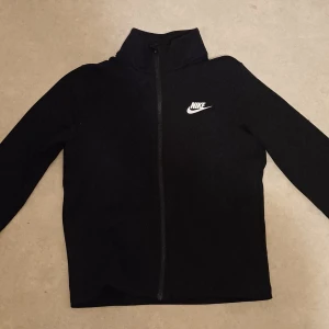 Svart zip-jacka från Nike - Svart jacka från Nike med hel dragkedja och hög krage. Stor Nike-logga på ryggen och mindre logga på bröstet. Jackan har lång ärm och är tillverkad i ett mjukt syntetmaterial. Perfekt för träning eller som en sportig vardagsjacka.