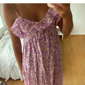 Lila blommig klänning med volang - Söt lila klänning med blommigt mönster och volangdetalj vid bysten. Klänningen har smala axelband och en luftig passform, perfekt för varma dagar.