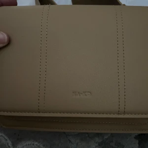 Beige axelväska från NA-KD - Säljer en stilren beige axelväska från NA-KD med bred axelrem och diskret logga. Väskan har flera fack, dragkedja och magnetknapp framtill. Perfekt för att bära det viktigaste med snygg och enkel design.