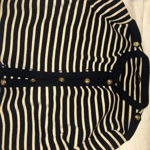 Svartvit randig kofta från Massimo Dutti - Säljer en svart och vit randig kofta från Massimo Dutti med dekorativa guldknappar framtill. Koftan är stickad och har långa ärmar samt rund halsringning. Perfekt för att lyfta din outfit med en klassisk touch.