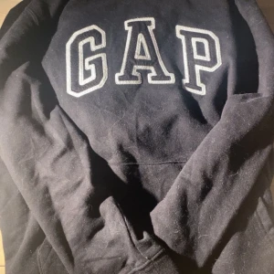 Svart hoodie från GAP - Svart hoodie från GAP med stor vit logga framtill. Klassisk modell med huva och magficka. Tillverkad i en mjuk bomulls- och polyestermix för extra komfort.