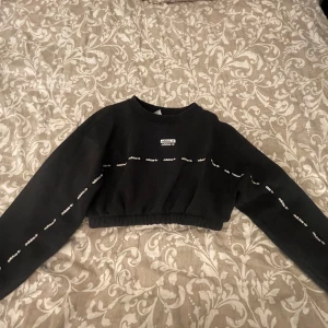 Svart croppad sweatshirt från Adidas - Säljer en svart croppad sweatshirt från Adidas med logga på bröstet och längs ärmarna. Tröjan har långa ärmar och resår i nederkanten för en snygg passform. Perfekt för en sportig och trendig look.