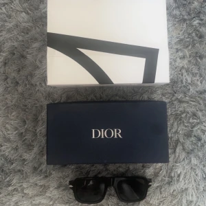 Svarta solglasögon DiorBlackSuit N21 från Dior - Snygga svarta solglasögon från Dior, modell DiorBlackSuit N21. Glasögonen har en markerad, fyrkantig form och är tillverkade i blankt svart acetat med diskreta silvriga detaljer och CD-logga på skalmen. Perfekt accessoar för en trendig look.